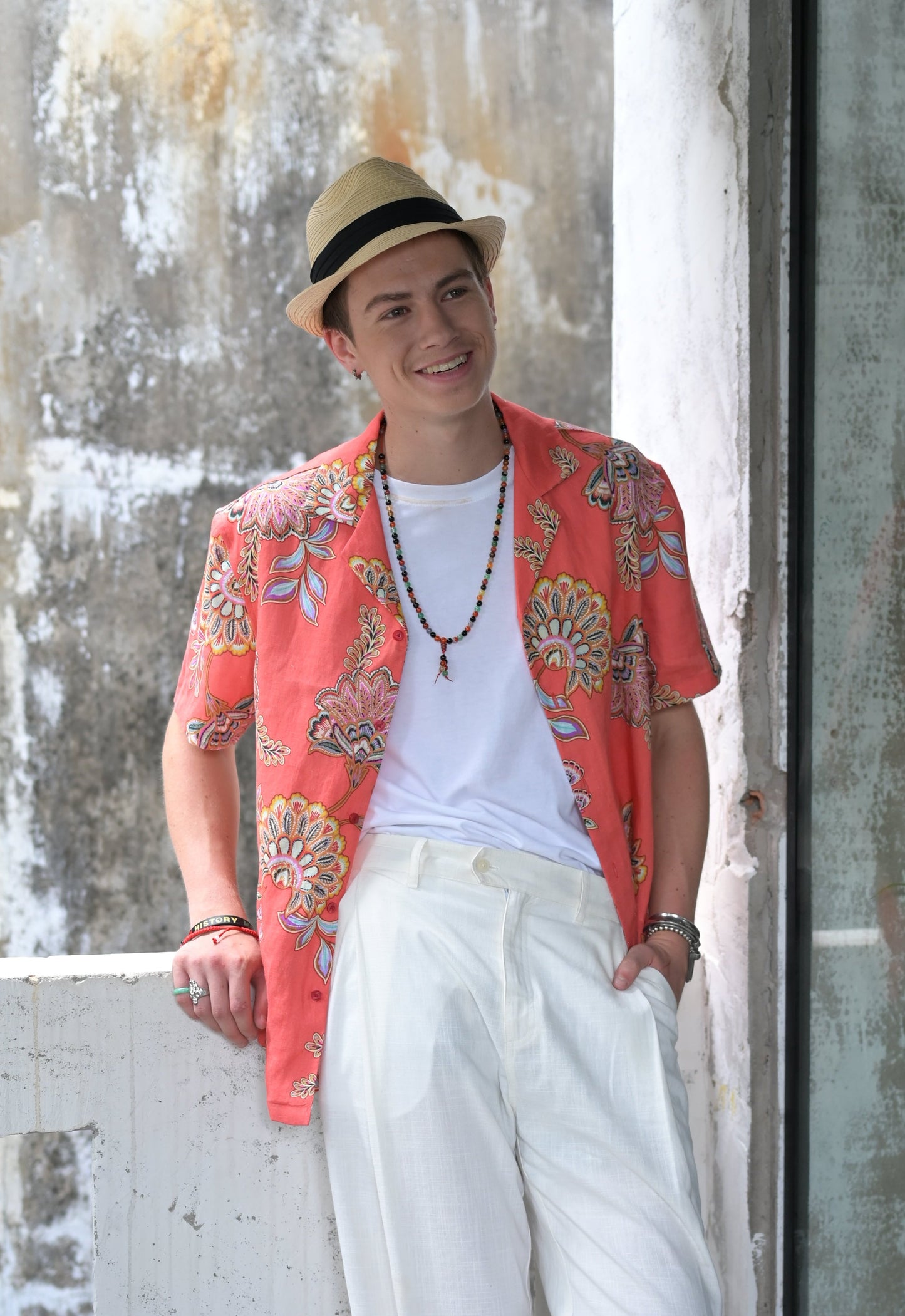 Man wearing coral coloured Hawaiian style shirt with floral print, white cotton tee and white linen pants standing at balcony