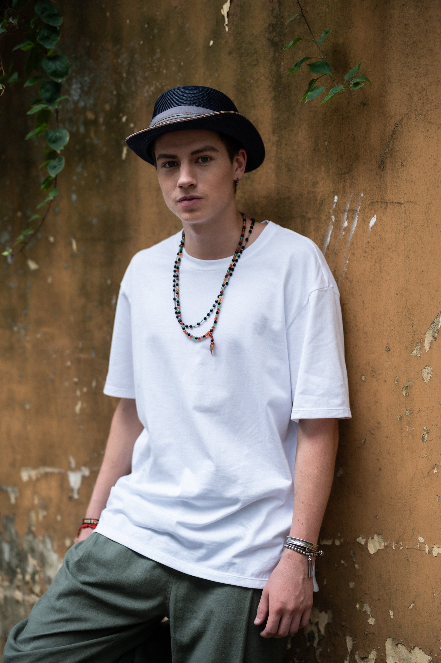 Person wearing a white t-shirt, green pants, and a black hat against a urban gardne wall.