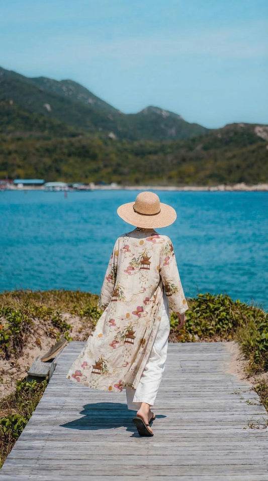 Woman walking along a beach boardwalk wearing a Saigon Concepts kimono-style robe with brown pagodas and pink Asia inspired swirls printed on a cream linen organza fabric - back view detail