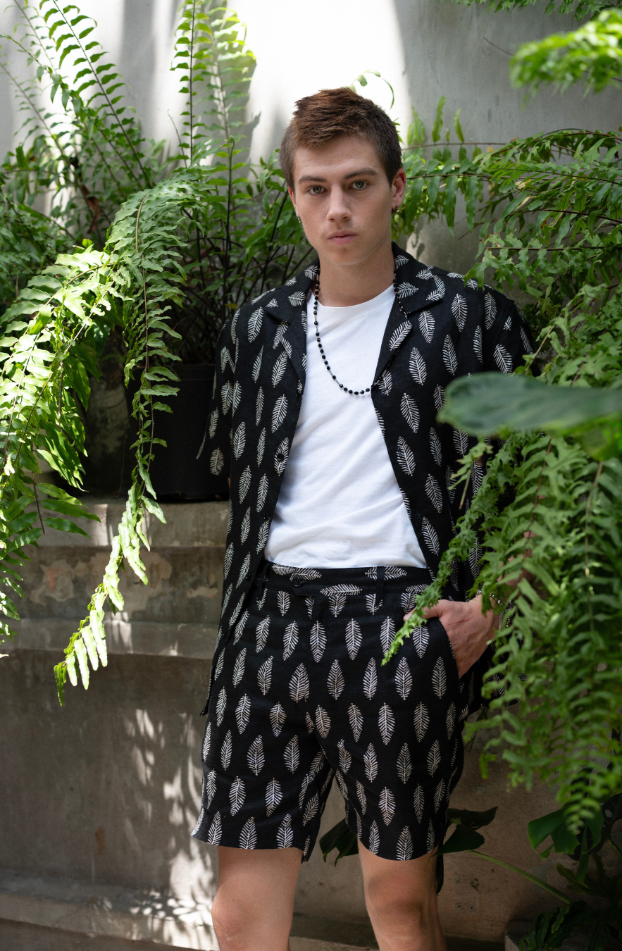 Young man wearing matching Hawaiian style shirt and shorts in black colour with white fern print standing in a garden