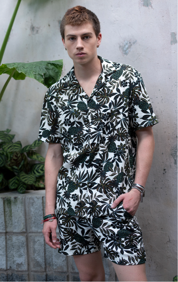 Man wearing matching black and white fern patterned Hawaiian shirt and shorts standing against a textured wall with plants.