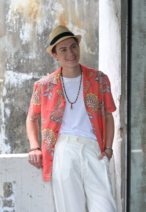 Man wearing coral coloured Hawaiian style shirt with floral print, white cotton tee and white linen pants standing at balcony