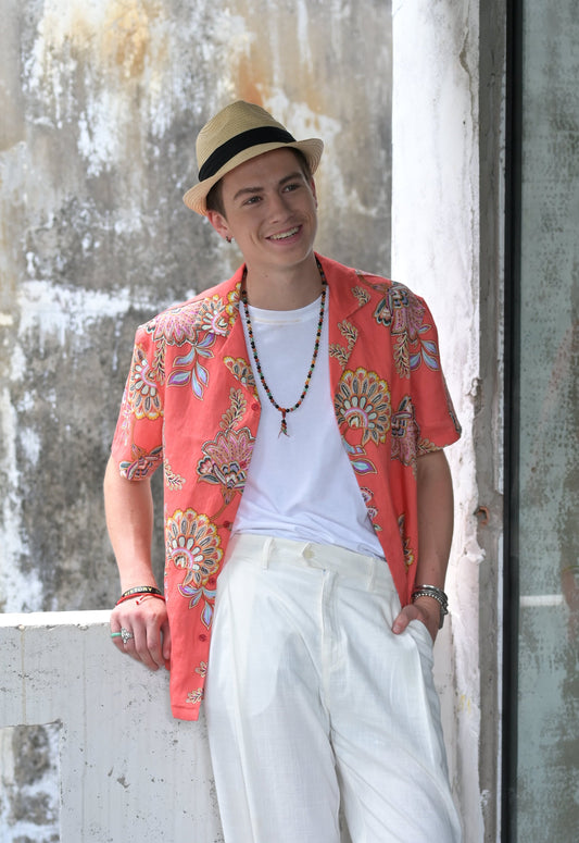 Man wearing coral coloured Hawaiian style shirt with floral print, white cotton tee and white linen pants standing at balcony