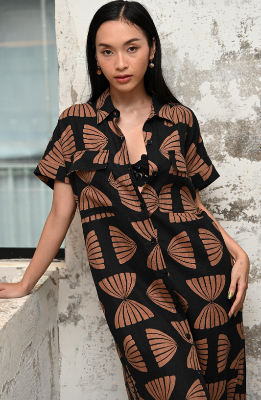 Woman wearing a black and copper maxi shirt dress with copper abstract pattern against a textured wall.