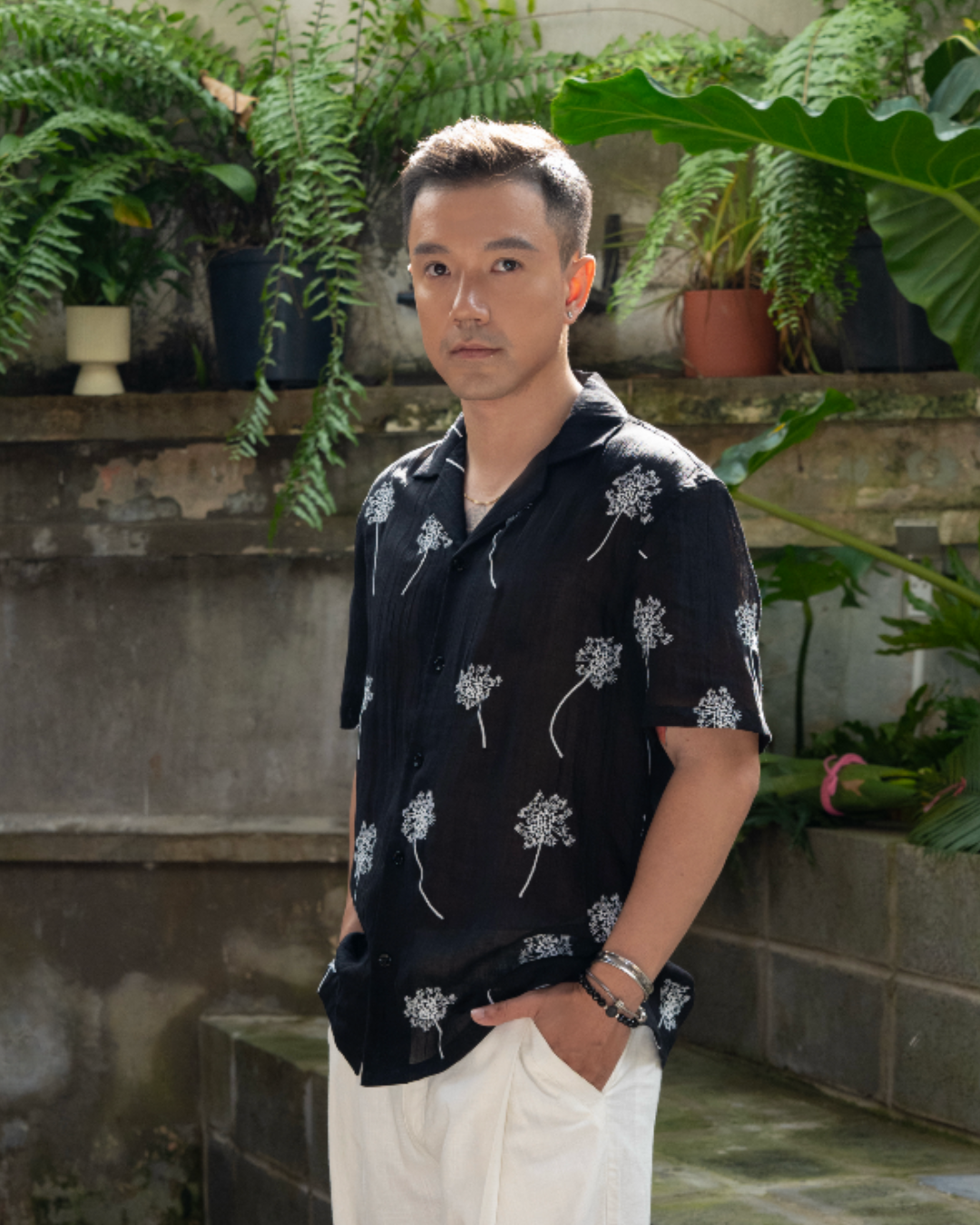 Man wearing a black linen shirt with white embroidered flowers standing in outdoor garden with plants in background