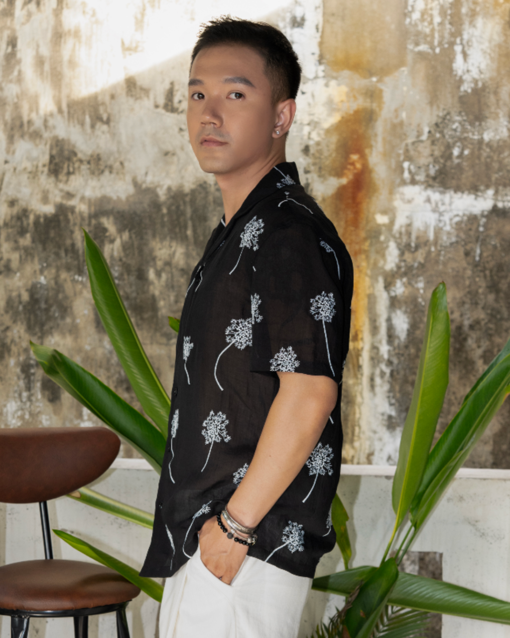 Man wearing a black linen shirt with white embroidered flowers standing in outdoor garden with plants in background