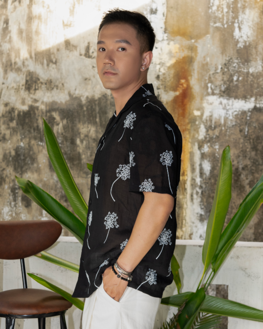 Man wearing a black linen shirt with white embroidered flowers standing in outdoor garden with plants in background