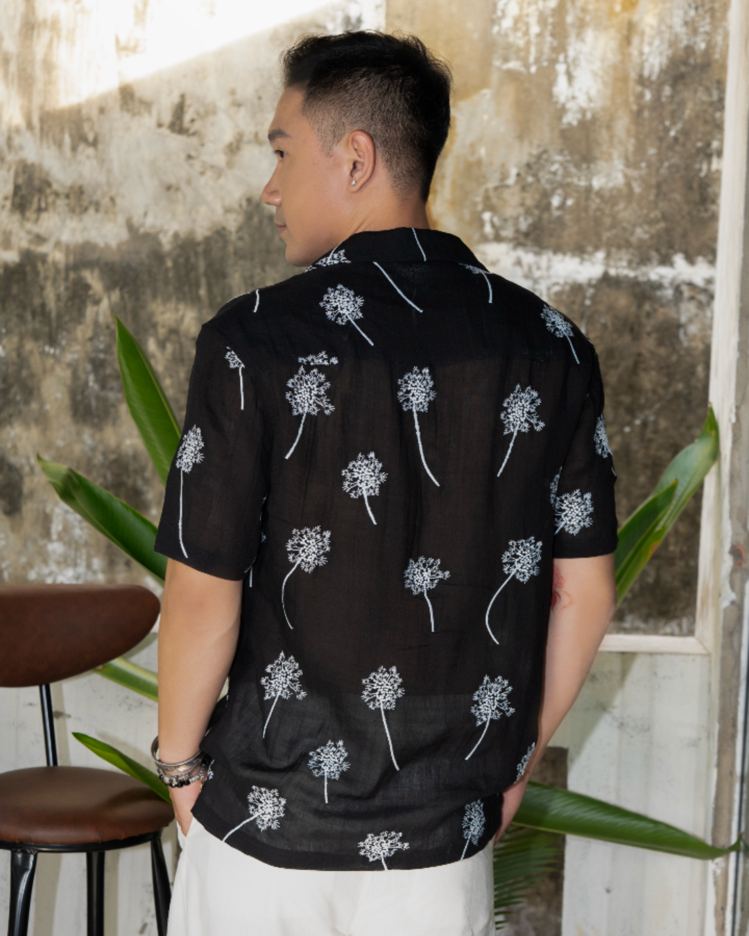 Man wearing a black linen shirt with white embroidered flowers standing in outdoor garden with plants in background