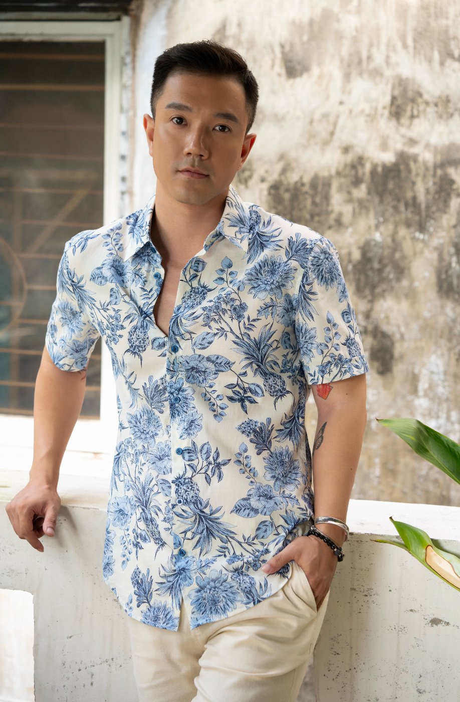 Man wearing a blue and white floral shirt and white shorts against a textured wall.