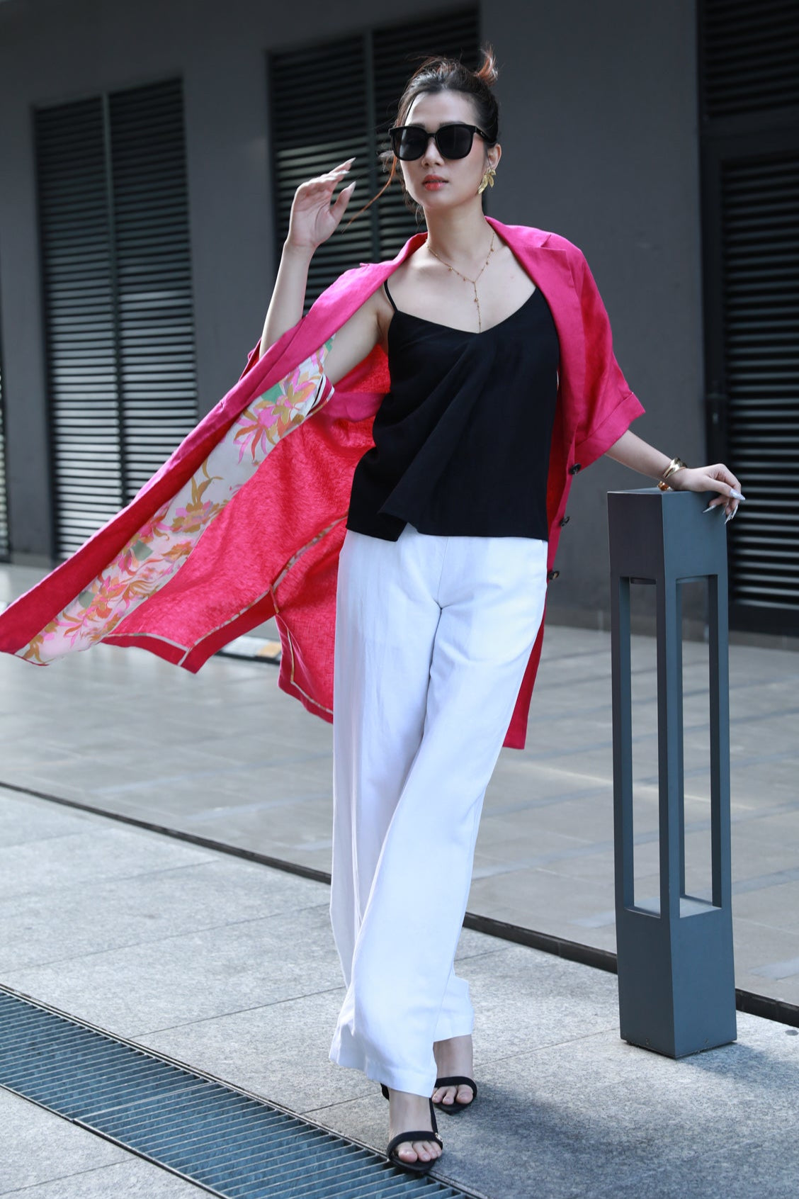 Woman in black top and white pants with a fuchsia pink linen coat on a sidewalk.