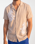 A man wearing a beige short-sleeve button-up shirt with white floral embroidery on the chest and lower front, paired with blue pants, against a plain light background.