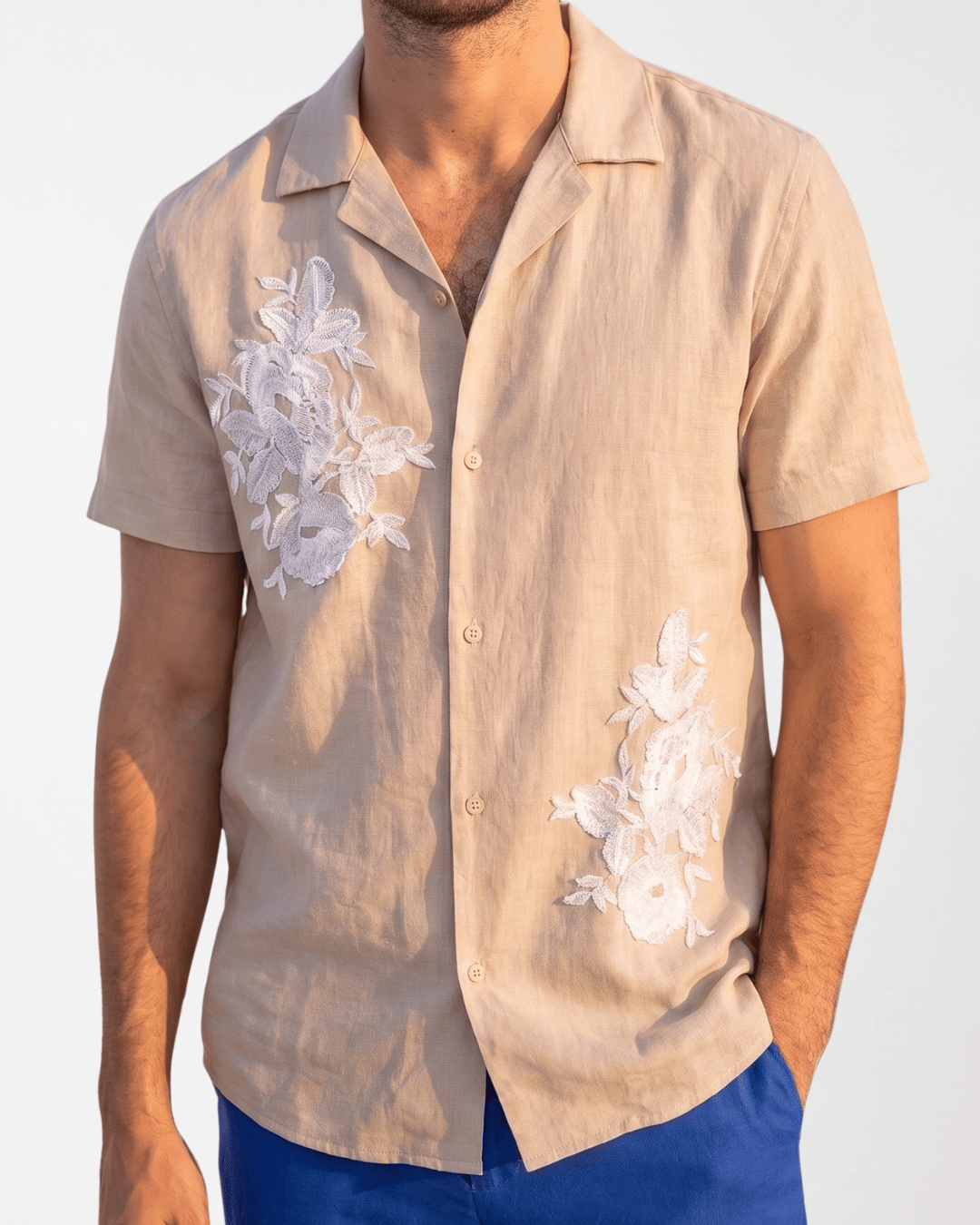 A man wearing a beige short-sleeve button-up shirt with white floral embroidery on the chest and lower front, paired with blue pants, against a plain light background.