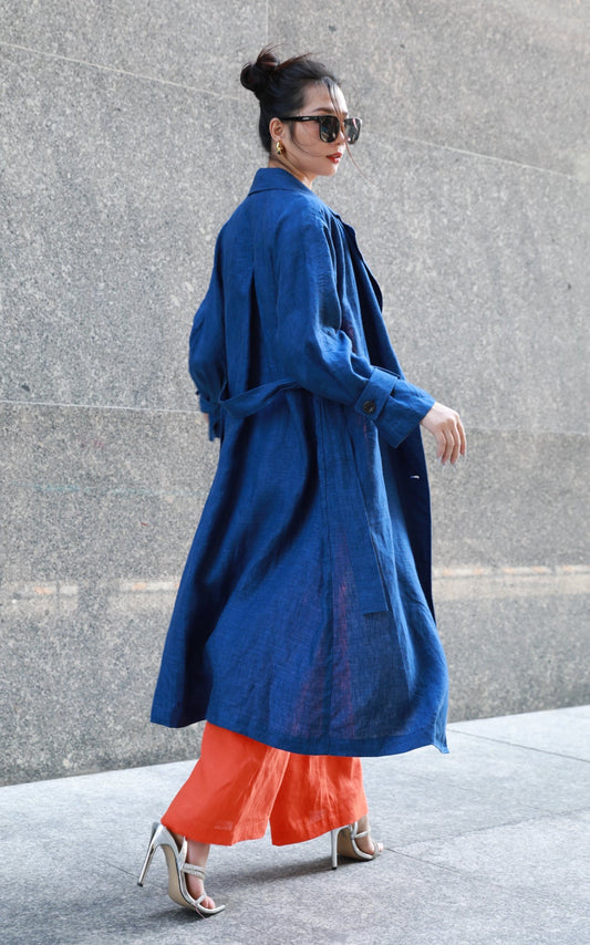Woman wearing a dark blue trench coat and orange pants against a gray wall.