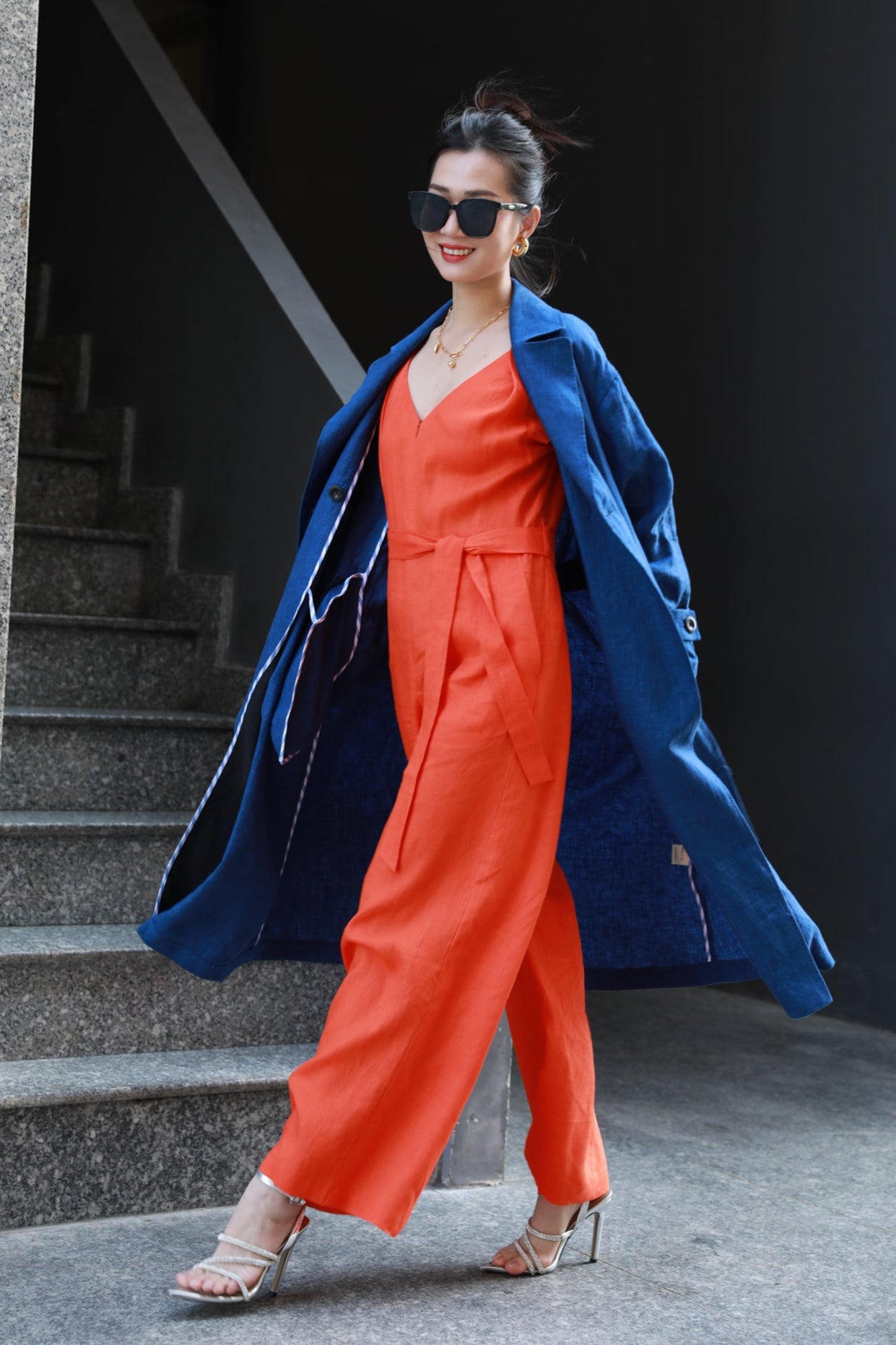 Woman in a dark blue trench coat and orange jumpsuit standing at a staircase.