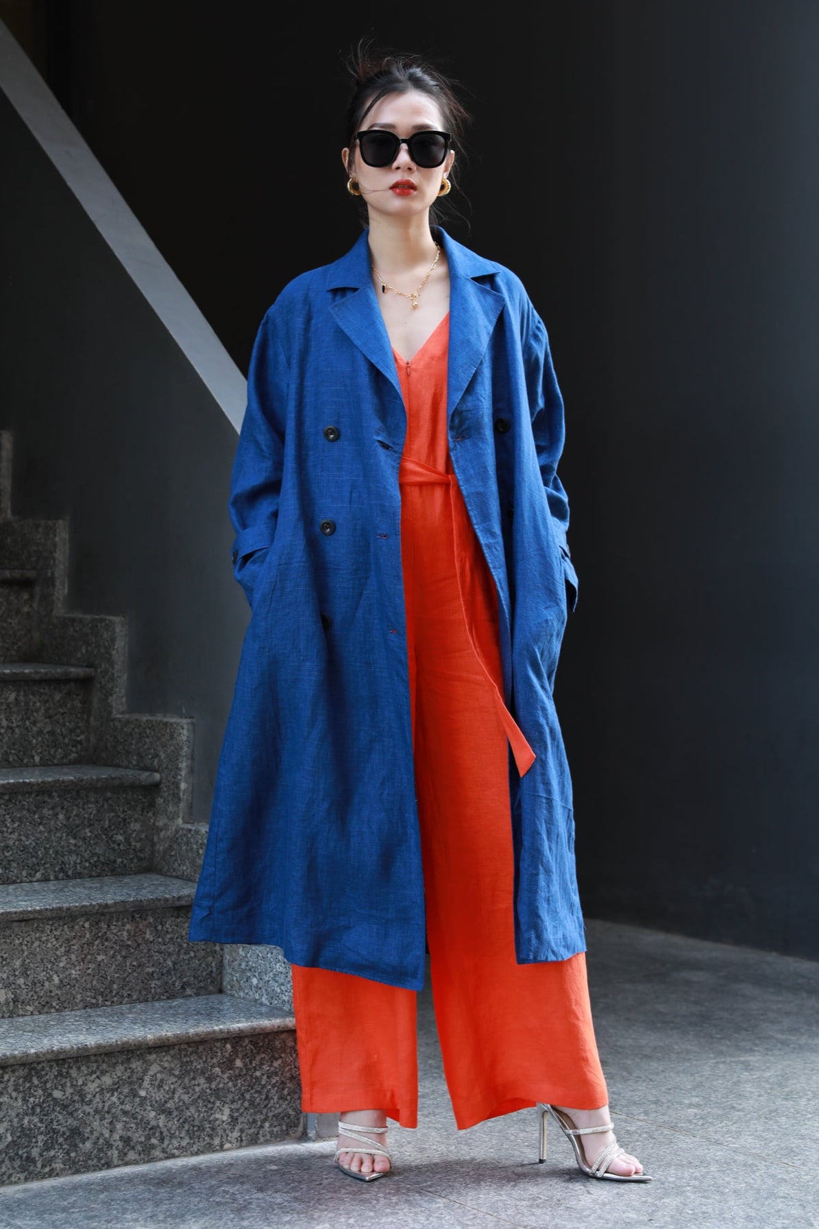 Woman wearing a navy blue trench coat over an orange jump suit standing at a staircase.