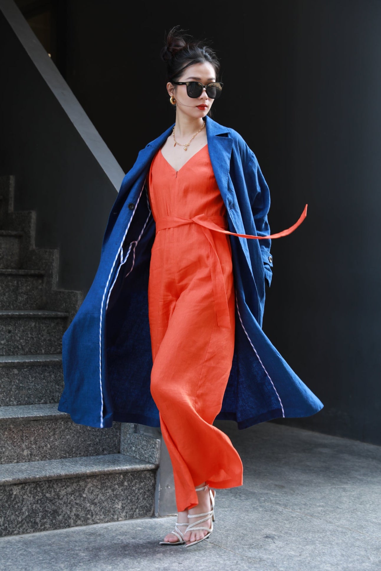 Woman in an orange jumpsuit and navy blue trench coat standing at a staircase.
