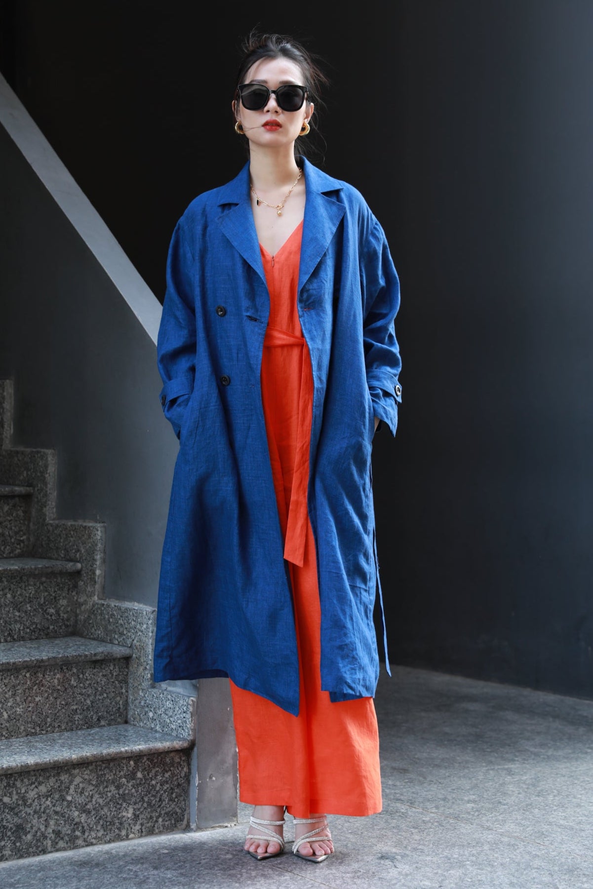 Woman wearing a navy blue trench coat over an orange jump suit standing at a staircase.