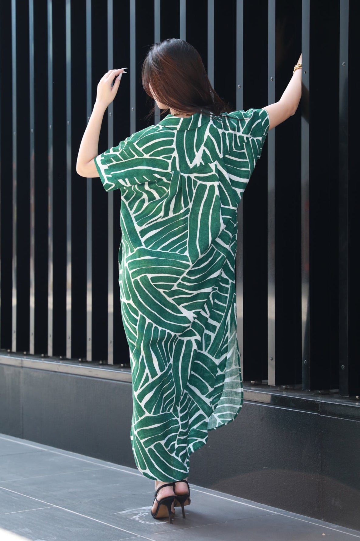 Woman wearing a green leaf-patterned dress against a black metal fence showing the back detail of the dress.