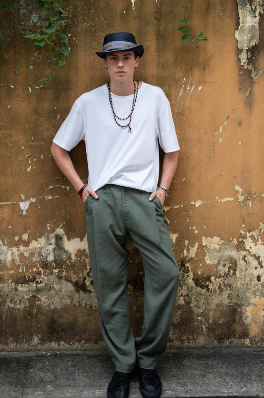 Person wearing a white t-shirt, green pants, and a black hat against a textured wall.