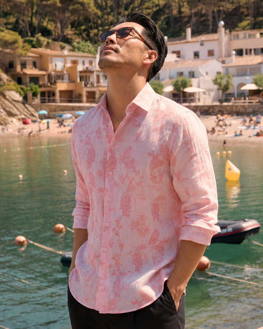 A man wearing a light pink long-sleeve button-up shirt with a darker pink abstract pattern, standing in front of a sunny beach and coastal town background.