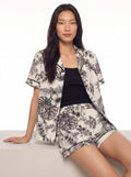 Female model seated in a cream and black botanical-print resort shirt and matching shorts against a white studio background