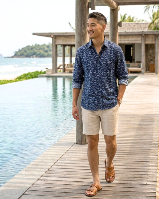 Man in a navy blue patterned long sleeve shirt and beige shorts standing on a wooden deck by a pool with a scenic asian background.
