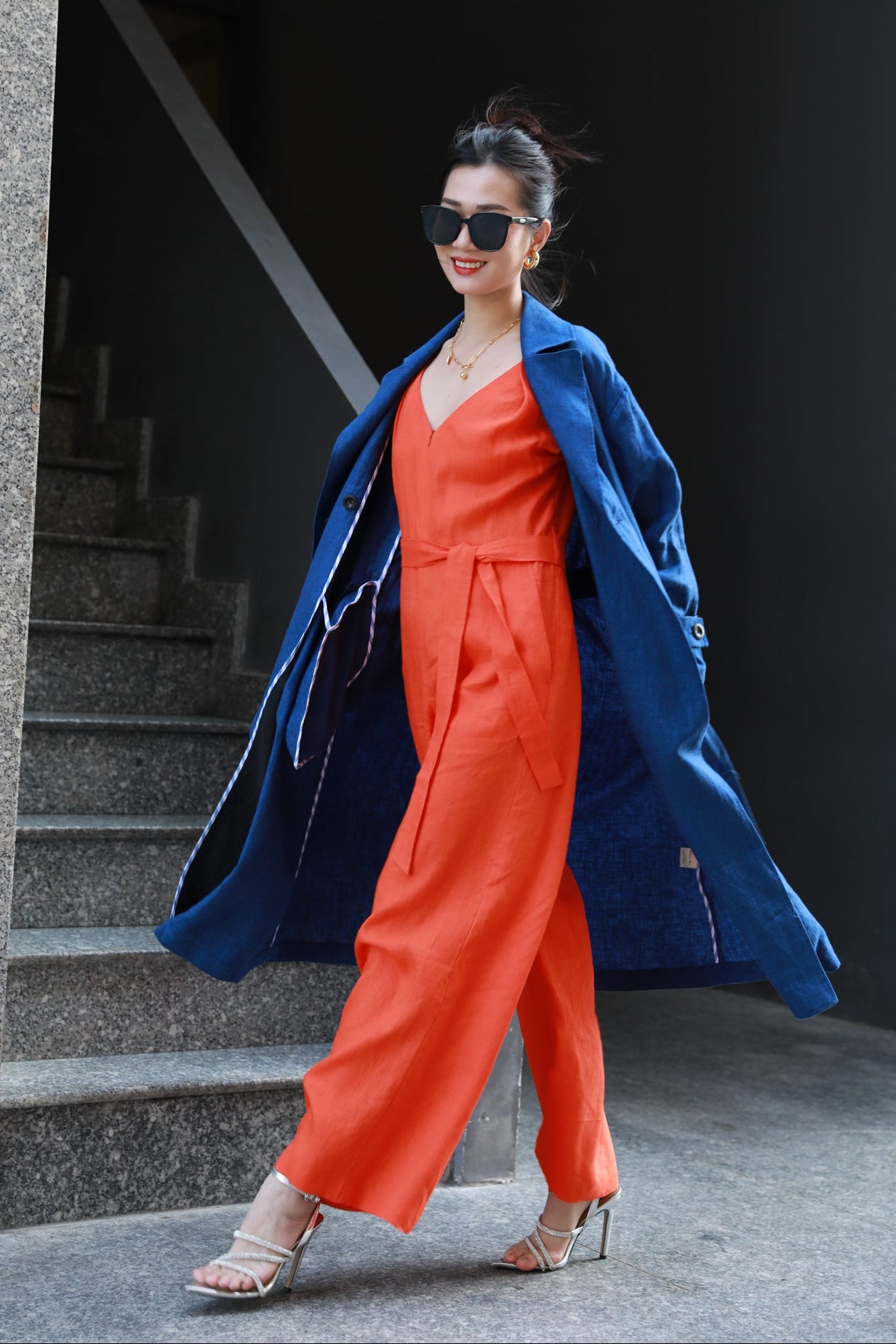 Woman in a Saigon Concepts blue linen trench coat and orange jumpsuit standing at a staircase.