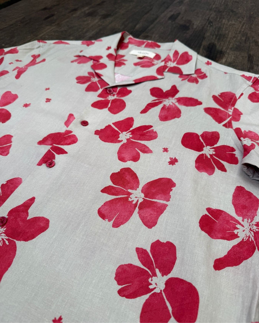 Beige-cream colour aloha style shirt with ruby red floral pattern on a wooden surface - lay flat side view