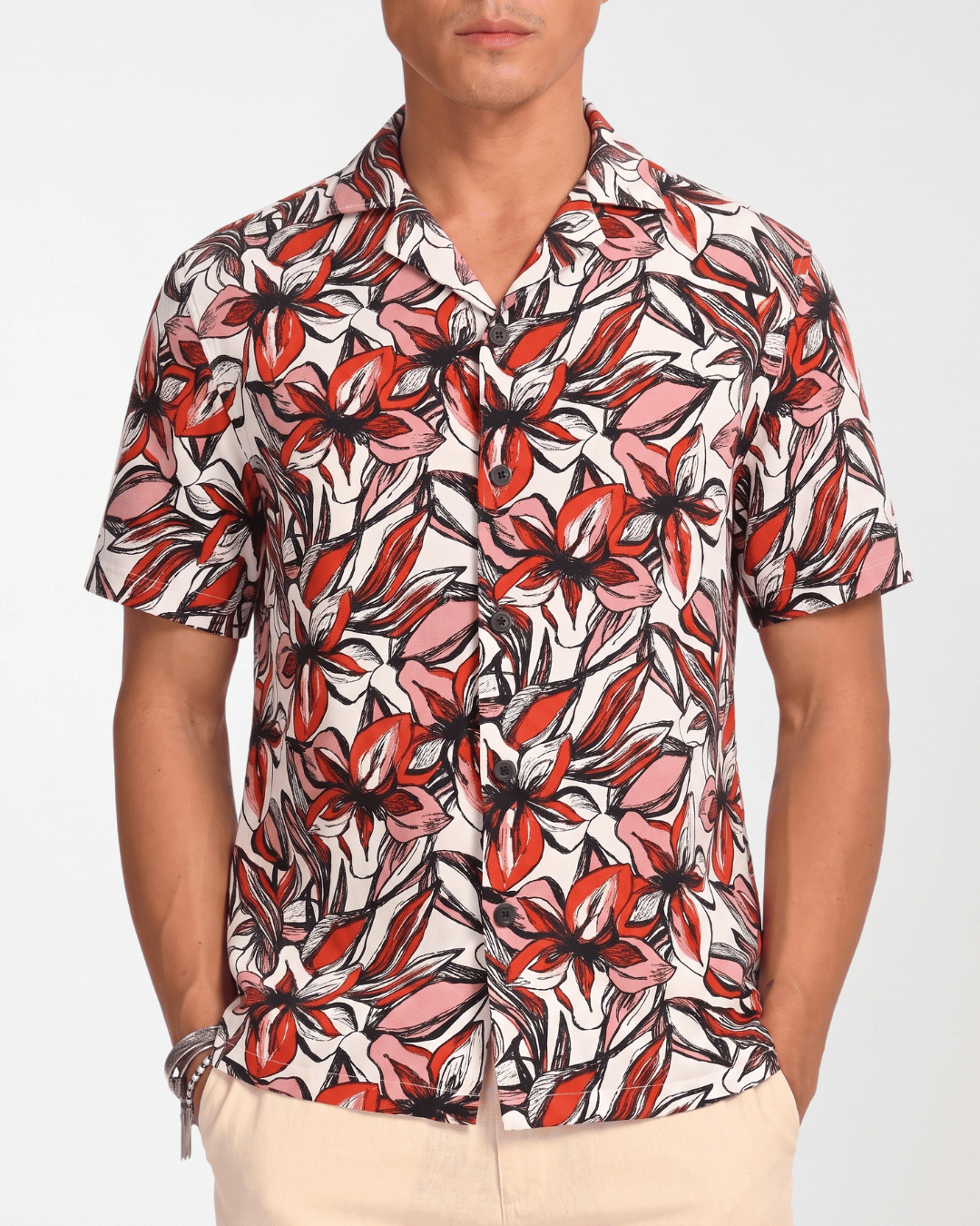 A man wearing a short-sleeve button-up camp shirt with a bold red, pink, and black tropical floral print on a white background, paired with beige pants.