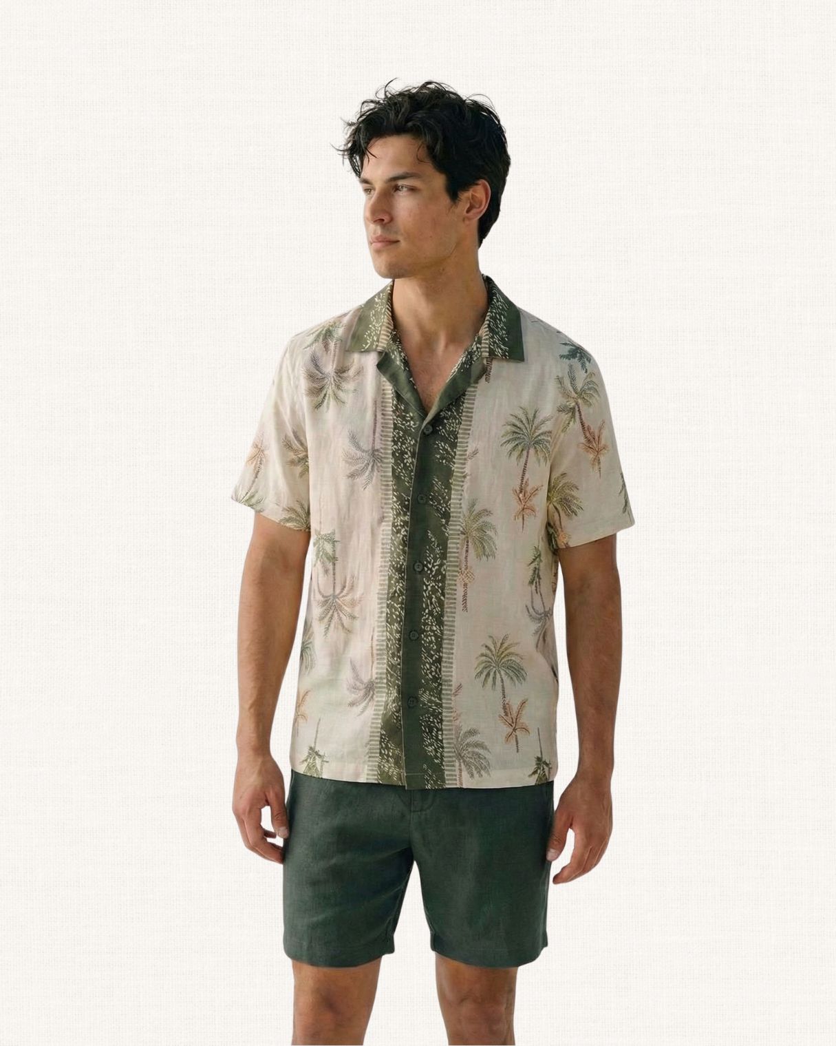 Man wearing a cream colour Hawaiian style shirt with palm tree pattern and dark green collar shirt and shorts against a white background 
