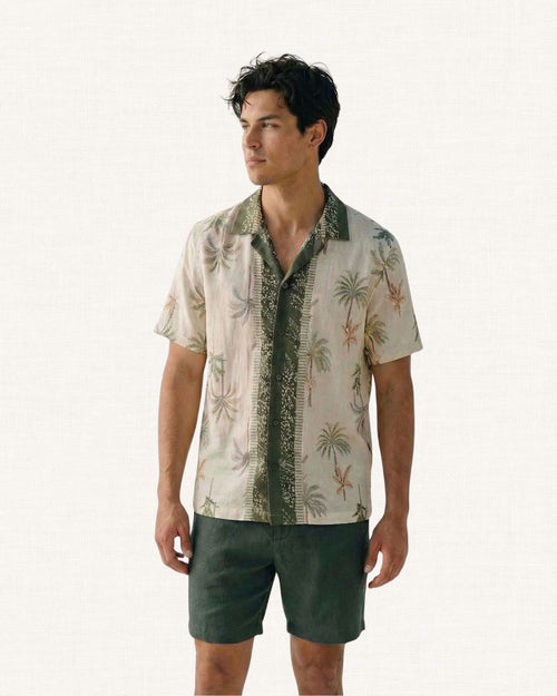 Man wearing a cream colour Hawaiian style shirt with palm tree pattern and dark green collar shirt and shorts against a white background 