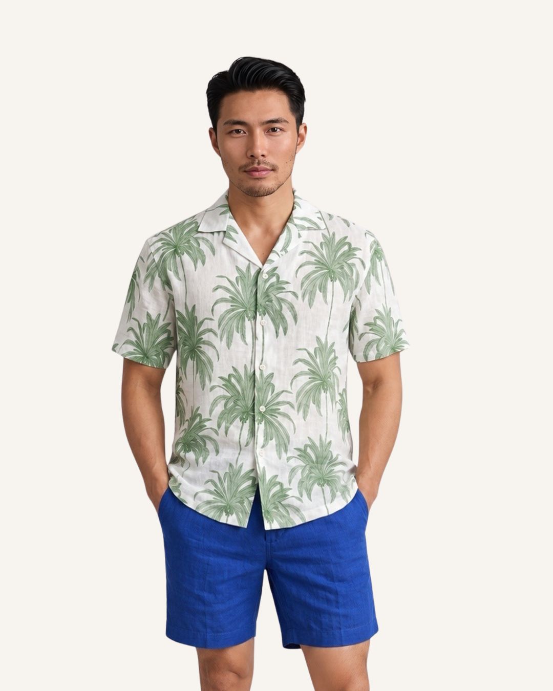 Male model standing against white background wearing Saigon Concepts Linen Hawaian /Aloha Short Sleeve Shirt with green palm print with cream background and cobalt blue linen shorts