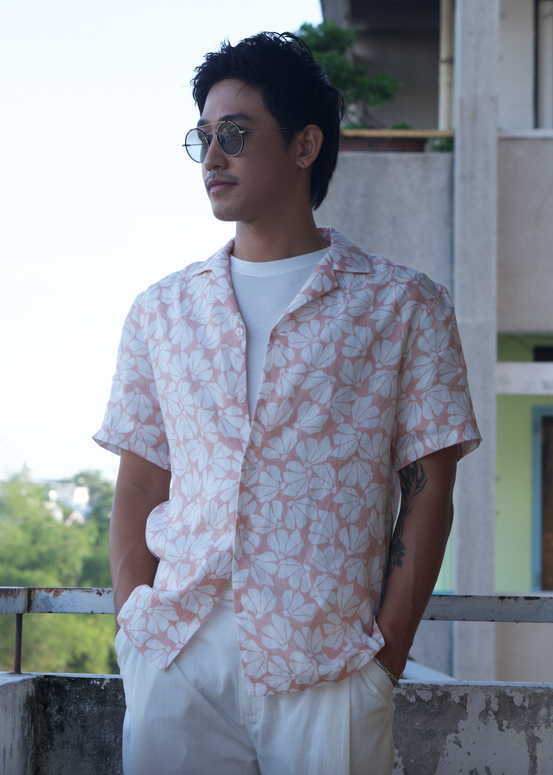 A man wearing a white petals linen Hawaii shirt with a pink floral pattern, paired with white trousers, standing on a balcony.