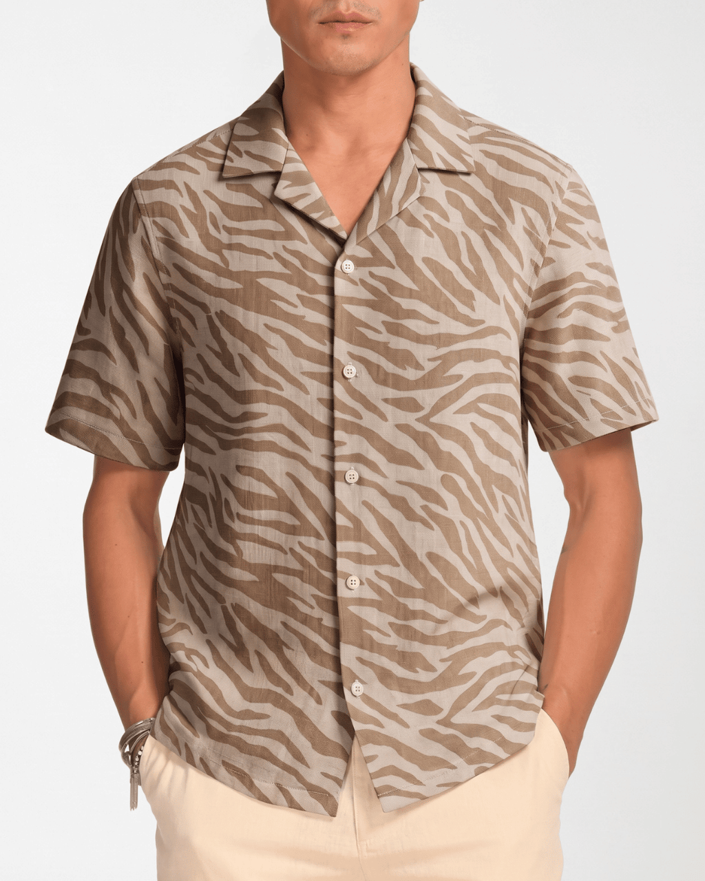 A man wearing a short-sleeve button-up shirt with a beige and brown zebra print, paired with light beige pants, against a plain light gray background.