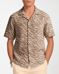 A man wearing a short-sleeve button-up shirt with a beige and brown zebra print, paired with light beige pants, against a plain light gray background.