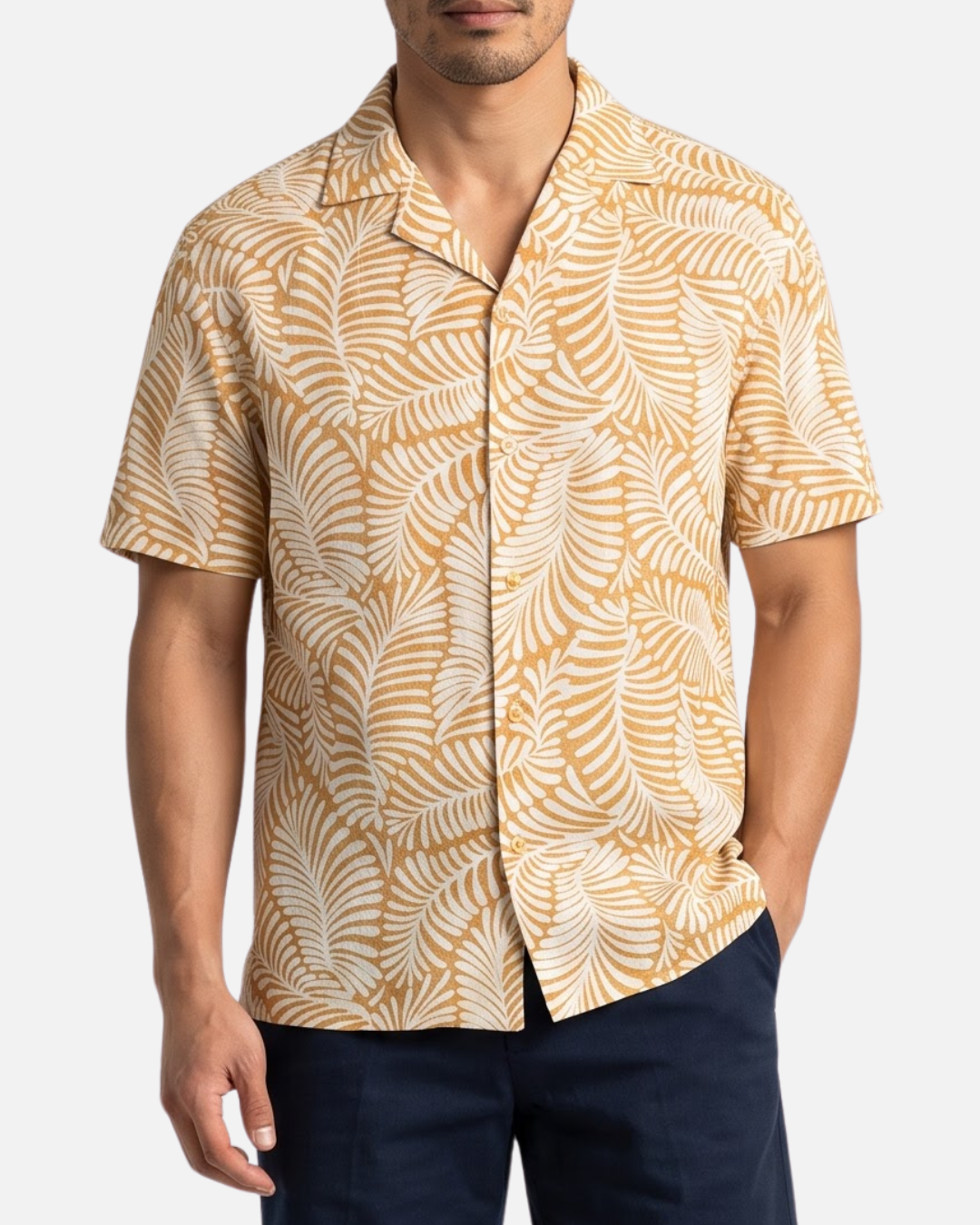 A man wearing a short-sleeve button-up camp shirt with a mustard yellow and white palm leaf pattern, paired with navy blue pants, against a light gray background.