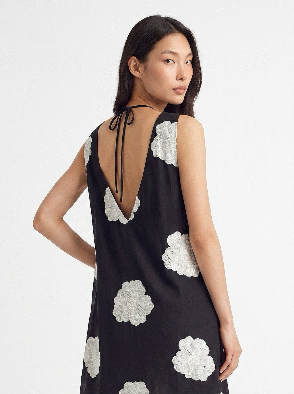 A woman shown from the back wearing a sleeveless black tunic top with large white floral applique and a tie-back neckline, paired with off-white wide-leg pants, against a plain light gray studio background.