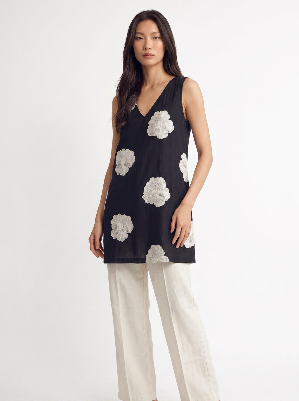 A woman modeling a sleeveless black tunic top with large white floral applique details, paired with off-white wide-leg trousers, against a plain white studio background.