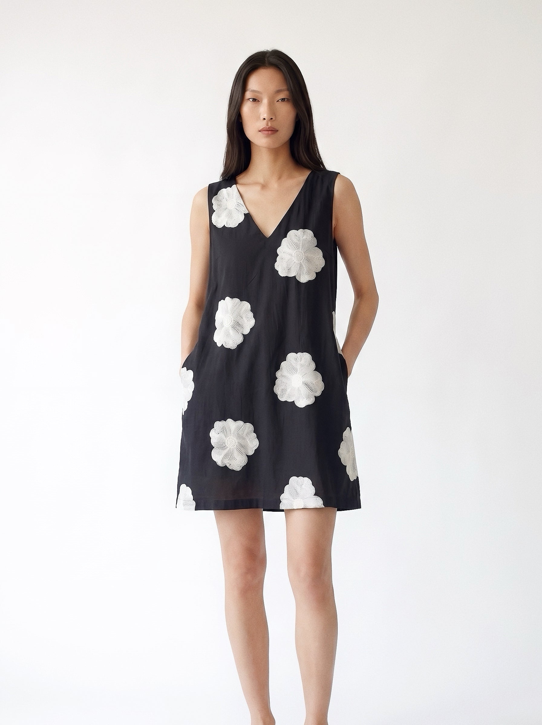 A woman modeling a sleeveless black V-neck mini dress with large white floral applique details, set against a plain white studio background.