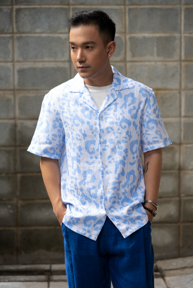 Man wearing a light blue leopard print Hawaiian style linen shirt and dark blue linen pants standing against a gray brick wall.
