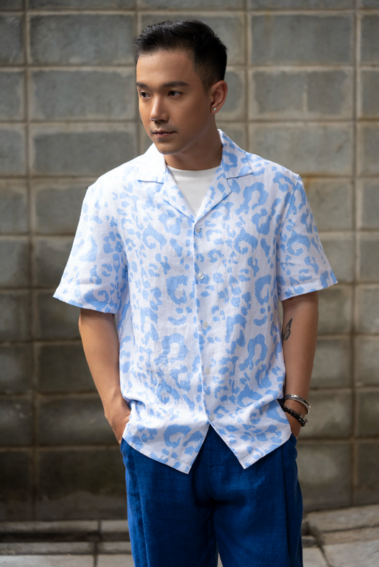 Man wearing a light blue leopard print Hawaiian style linen shirt and dark blue linen pants standing against a gray brick wall.