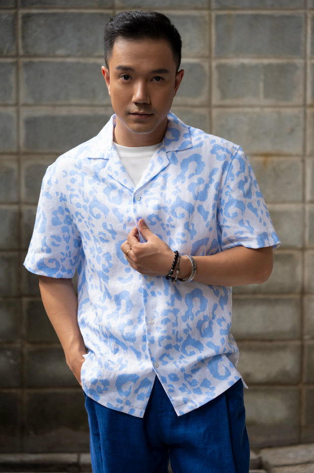 Man wearing a light blue leopard print Hawaiian style linen shirt and dark blue linen pants standing against a gray brick wall.