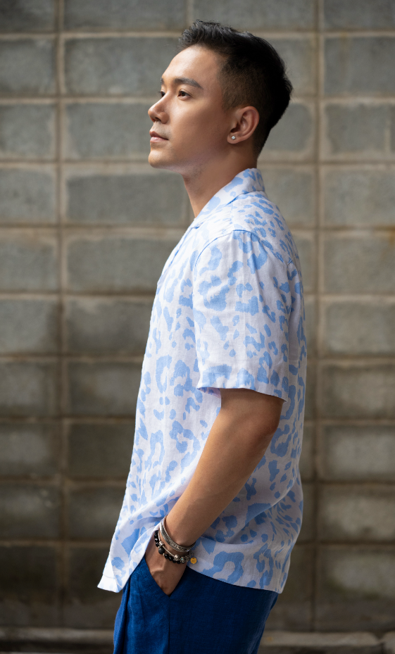 Man wearing a light blue leopard print Hawaiian style linen shirt and dark blue linen pants standing against a gray brick wall.