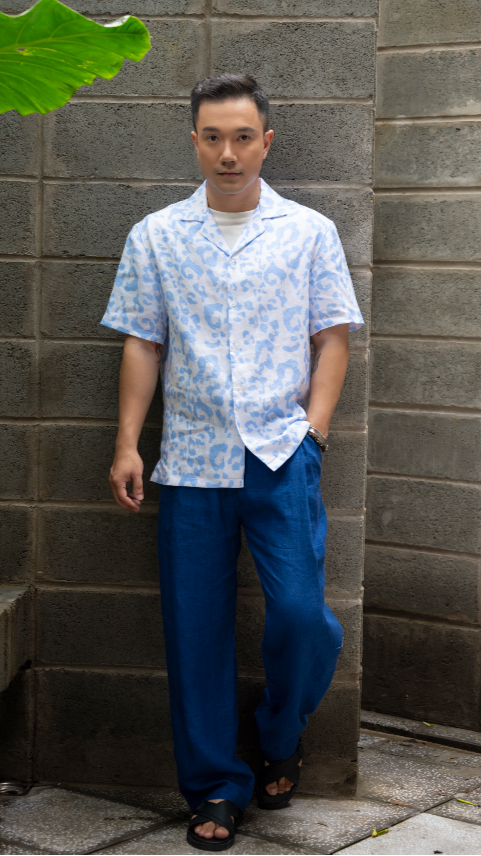 Man wearing a light blue leopard print Hawaiian style linen shirt and dark blue linen pants standing against a gray brick wall.