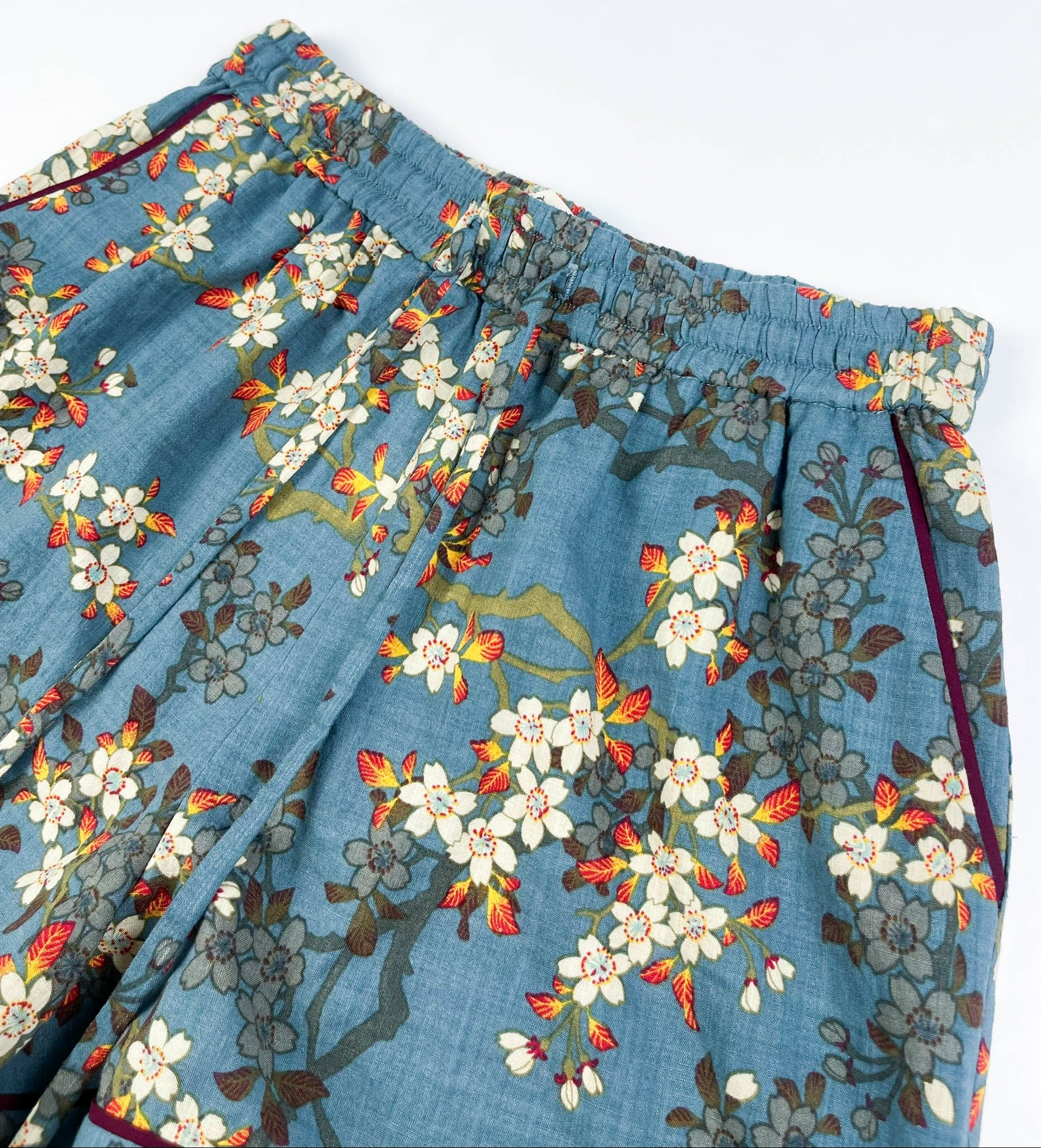 Female Lounge Shorts in teal blue with white blossom floral print. Flat front view of elasticated waistband, drawstring cord and side pockets with burgundy contrast piping on seams - Saigon Concepts