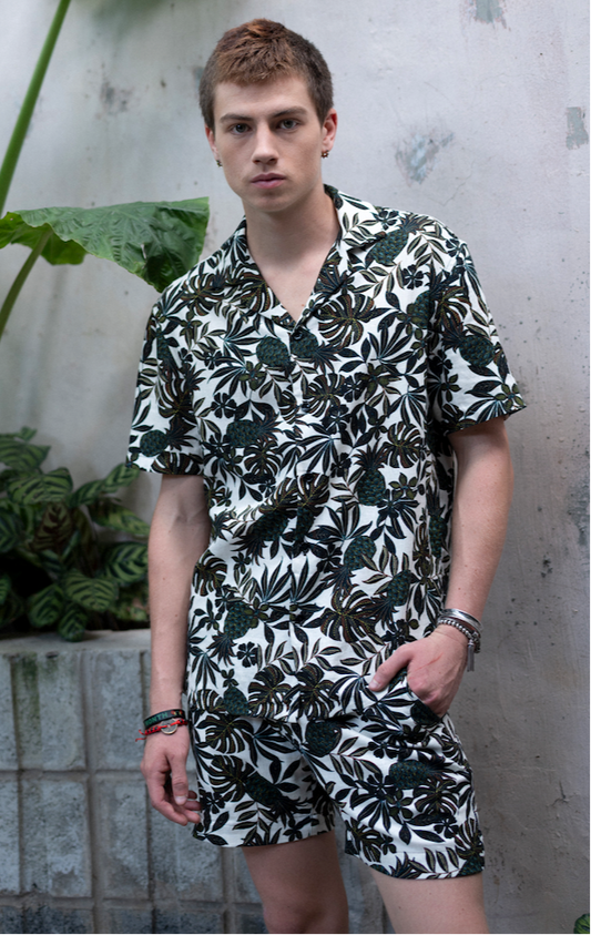 Man wearing matching black and white fern patterned Hawaiian shirt and shorts standing against a textured wall with plants.