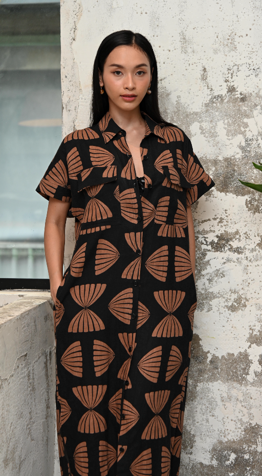 Woman wearing a black and copper maxi shirt dress with copper abstract pattern against a textured wall.