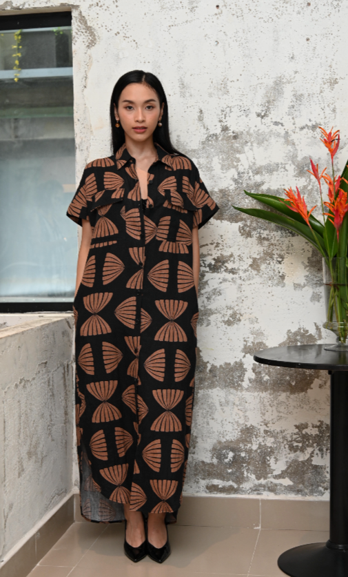 Woman wearing a black and copper maxi shirt dress with copper abstract pattern against a textured wall - full length image.
