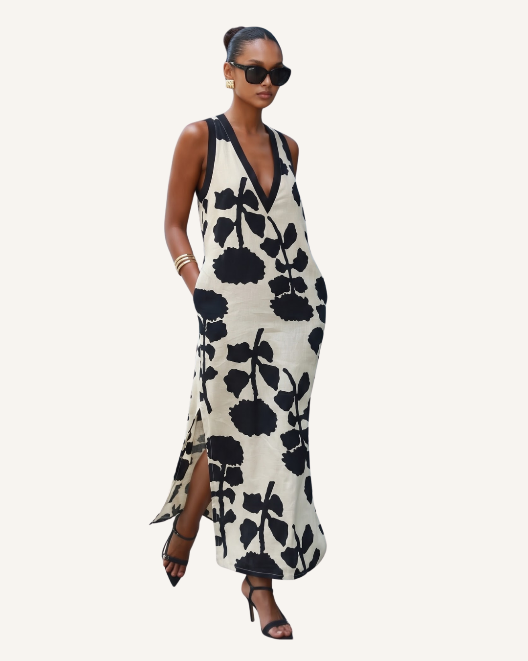 A woman modeling a sleeveless, deep V-neck maxi dress with a black floral ikat pattern on a cream background, styled with black sunglasses, bracelets, and strappy high-heeled sandals against a solid off-white background.
