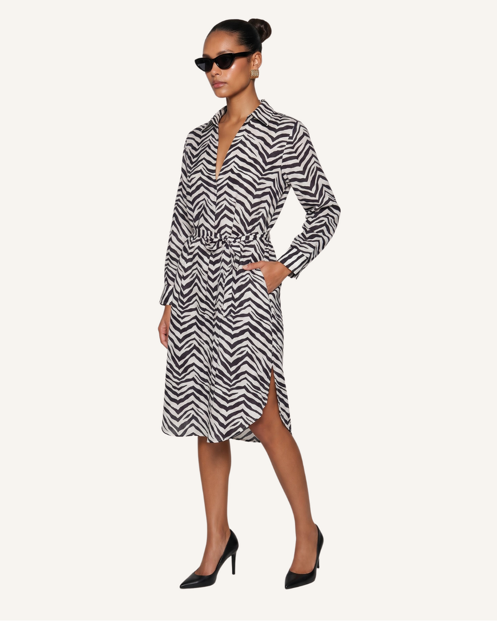 A woman modeling a long-sleeve midi dress with a black and white chevron pattern, a split V-neckline, and a matching waist tie, styled with black sunglasses and black high heels against a solid off-white background.